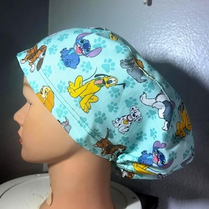 Może przedstawiać: Jasnoniebieski czepek chirurgiczny z wzorem postaci z kreskówek i odcisków łap. Czepek przedstawia postacie takie jak Stitch, Pluto i dalmatyńczyk. Wzór powtarza się na tkaninie, tworząc zabawną i kolorową ozdobę.