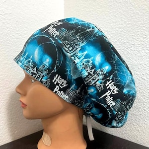 May include: A blue and black patterned scrub cap featuring the text "Harry Potter" and illustrations of Hogwarts. The cap has a white tie in the back for adjustment. The cap is on a mannequin head.