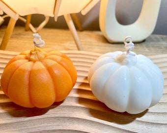 Puede incluir: Dos velas con forma de calabaza, una naranja y otra blanca, sobre una superficie de madera.