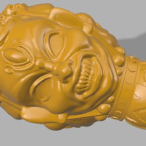 May include: A gold-colored decorative object with a detailed, stylized face and a textured, patterned body. The face has a fierce expression with a wide mouth and sharp teeth.