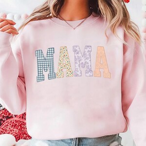 Könnte beinhalten: Hellrosa Sweatshirt mit dem Wort "MAMA" in dekorativen Buchstaben. Die Buchstaben sind gemustert: blau-weiß kariert für das erste "M", floral für das "A", lila floral für das zweite "M" und pfirsichfarben für das letzte "A".