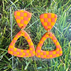 May include: A pair of dangle earrings featuring a checkered pattern in vibrant orange and yellow. Each earring consists of a small trapezoidal top piece connected by a gold-toned metal ring to a larger, hollow triangular bottom pendant.