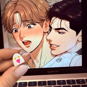 May include: A small white ghost-shaped pin with two yellow circular eyes and a pink triangular mouth. The pin is held in front of a laptop screen displaying a digital illustration of two characters with expressive faces and the text TAP.