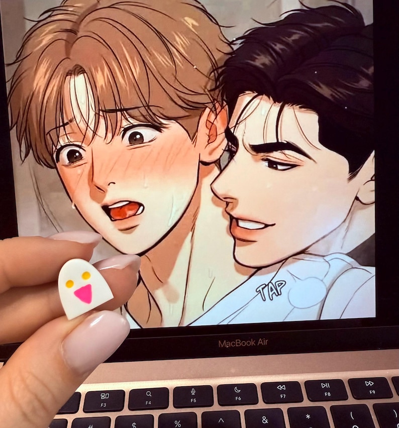May include: A small white ghost-shaped pin with two yellow circular eyes and a pink triangular mouth. The pin is held in front of a laptop screen displaying a digital illustration of two characters with expressive faces and the text TAP.