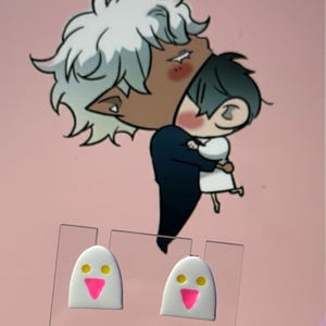 May include: A pair of white stud earrings shaped like ghosts with yellow circular eyes and a bright pink triangular mouth. The earrings are displayed on a clear plastic card against a background featuring an illustration of two characters embracing.