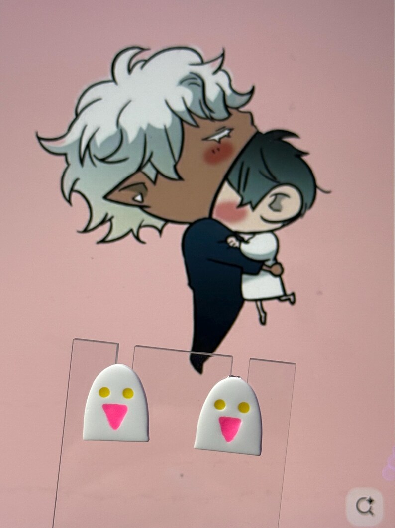 May include: A pair of white stud earrings shaped like ghosts with yellow circular eyes and a bright pink triangular mouth. The earrings are displayed on a clear plastic card against a background featuring an illustration of two characters embracing.