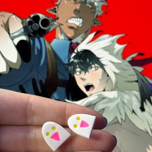 May include: A pair of white stud earrings shaped like rounded ghosts, each featuring two small yellow circular eyes and a bright pink triangular mouth, presented as a set of two individual pieces with a smooth, flat surface finish.
