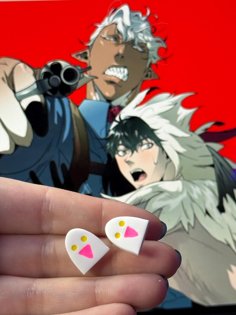 May include: A pair of white stud earrings shaped like rounded ghosts, each featuring two small yellow circular eyes and a bright pink triangular mouth, presented as a set of two individual pieces with a smooth, flat surface finish.
