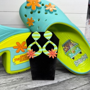 May include: A pair of green and white striped earrings with orange flower accents on a black display stand. The earrings are in front of a pair of teal clogs with orange flower details and a yellow sole with a Scooby-Doo graphic.