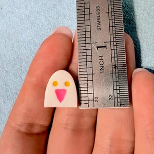 May include: A small, off-white, dome-shaped object featuring two yellow circular eyes and a pink triangular nose, positioned next to a stainless steel ruler that shows the object is approximately 0.5 inches in height.