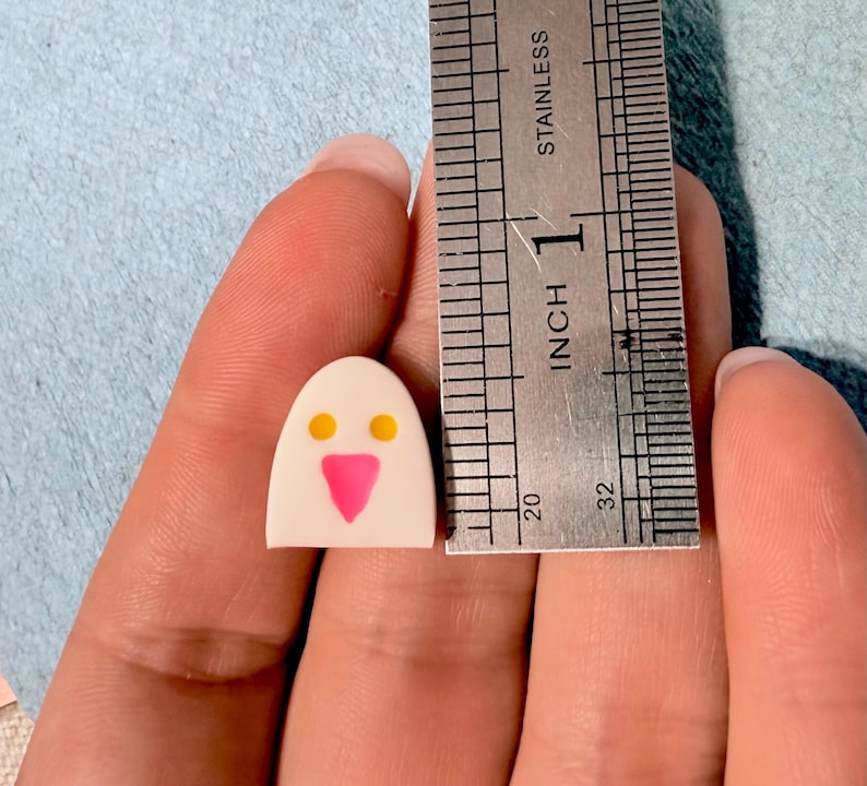 May include: A small, off-white, dome-shaped object featuring two yellow circular eyes and a pink triangular nose, positioned next to a stainless steel ruler that shows the object is approximately 0.5 inches in height.
