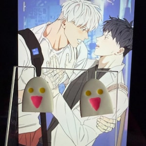 May include: A pair of white, dome-shaped stud earrings featuring a simple face design with two small yellow circular eyes and a pink triangular nose. The earrings are displayed on a clear acrylic stand in front of an illustration of two characters.