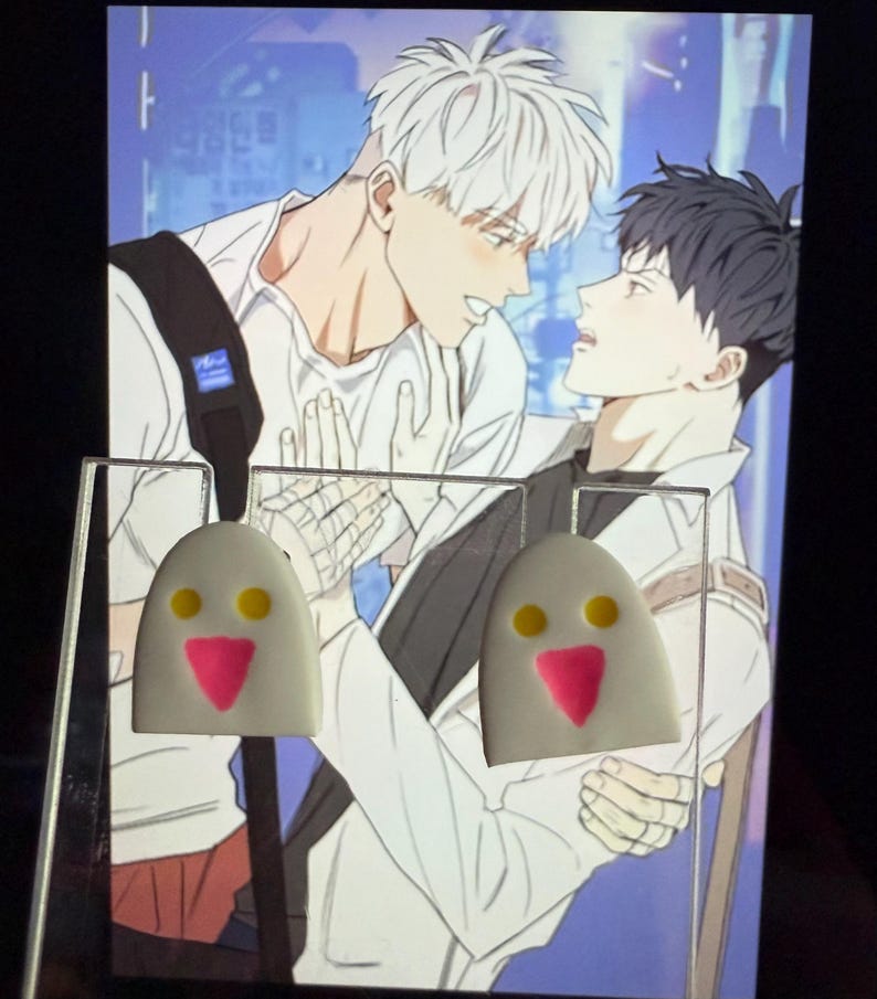 May include: A pair of white, dome-shaped stud earrings featuring a simple face design with two small yellow circular eyes and a pink triangular nose. The earrings are displayed on a clear acrylic stand in front of an illustration of two characters.