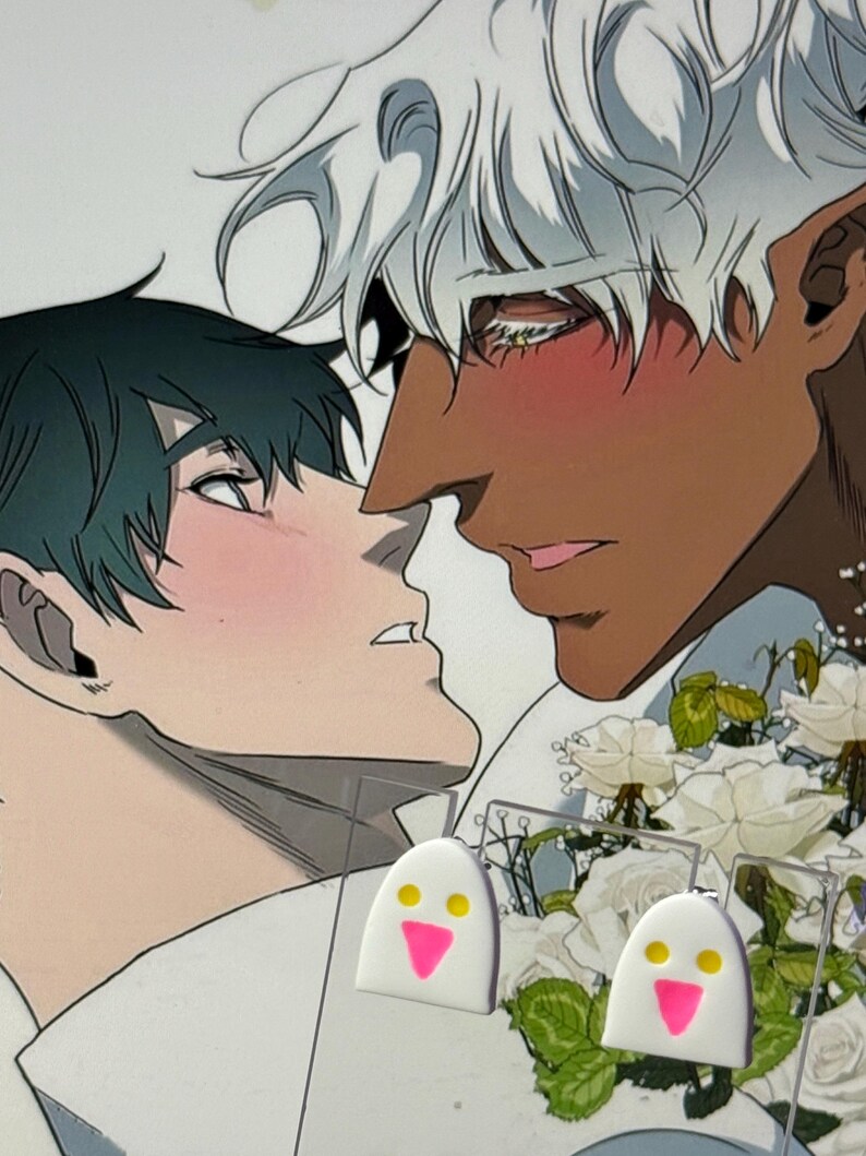 May include: A pair of white ghost-shaped earrings featuring two yellow circular eyes and a pink triangular mouth, displayed on a clear backing card in front of an illustration of two characters with white roses.