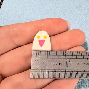 May include: A small, white, dome-shaped object with two yellow circular eyes and a pink triangular nose, positioned against a stainless steel ruler showing a width of approximately 0.5 inches. The object has a smooth, flat base and rounded top.