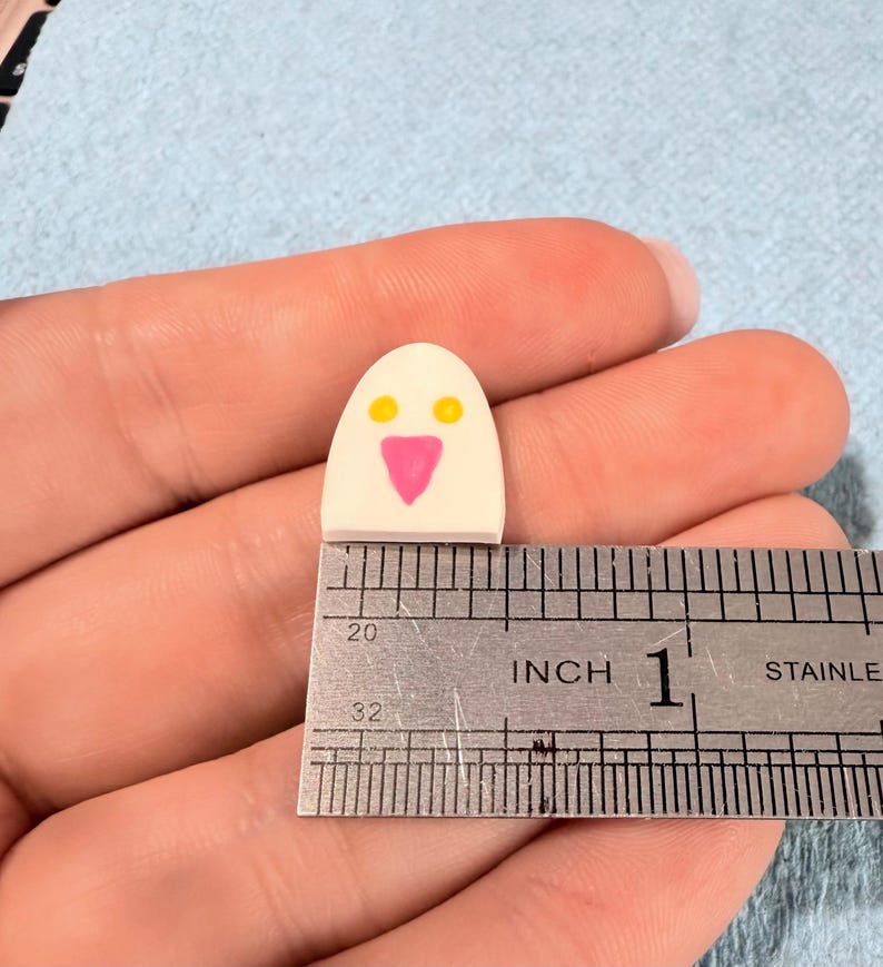 May include: A small, white, dome-shaped object with two yellow circular eyes and a pink triangular nose, positioned against a stainless steel ruler showing a width of approximately 0.5 inches. The object has a smooth, flat base and rounded top.