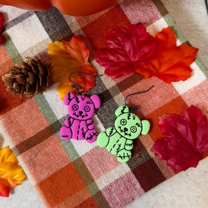 May include: A pair of Halloween-themed earrings featuring stitched teddy bear charms. One charm is pink and the other is green. The earrings are displayed on a plaid fabric background.