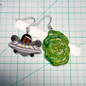 May include: A pair of handmade earrings. One earring is a silver UFO with black and yellow accents. The other earring is a green, swirled portal design. Both earrings have silver-colored ear wires.
