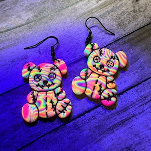 May include: A pair of colorful teddy bear earrings. Each bear is made of swirled neon colors with black stitching details and black ear wires. The bears have stitched mouths and button eyes.