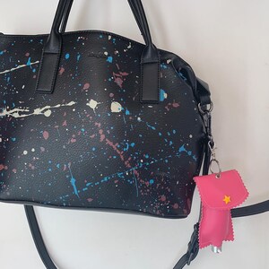Könnte beinhalten: Eine schwarze Tragetasche mit einem Sprühfarbendesign und einem rosa Handdesinfektionsmittelhalter mit einem gelben Stern, der an der Tasche befestigt ist.