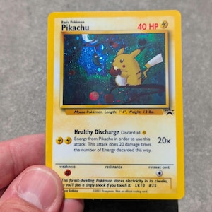 Op de afbeelding: Een gele Pikachu Pokémon-verzamelkaart. De kaart toont een illustratie van Pikachu met de tekst "Pikachu" en "40 HP". De tekst beschrijft de aanval "Gezonde Ontlading" en andere speld details. De kaart is 41 cm lang en weegt 5,9 kg.