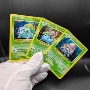 May include: Three Pokémon trading cards featuring Bulbasaur, Ivysaur, and Venusaur. The cards have a green and yellow border with character illustrations and text. The cards are held by a gloved hand.