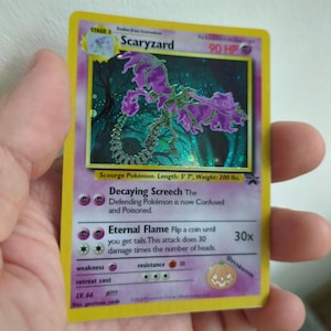 Könnte beinhalten: Eine Pokémon-Sammelkarte mit "Scaryzard". Die Karte hat einen gelben Rand und zeigt eine lila-grüne Monsterillustration. Der Text enthält "Decaying Screech" und "Eternal Flame". Die Karte wird in einer Hand gehalten.