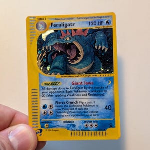 Könnte beinhalten: Eine Feraligatr Pokémon-Karte mit gelbem Hintergrund. Die Karte zeigt eine blaue Feraligatr-Illustration, den Namen "Feraligatr" und "120 HP". Text beschreibt die Fähigkeiten und Angriffe des Pokémon, typisch für eine Sammelkarte.