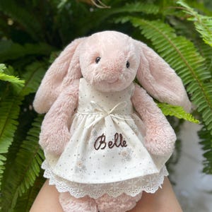 Könnte beinhalten: Ein rosa Plüschhase, der ein weißes Kleid mit floralem Muster und Spitzenbesatz trägt. Auf dem Kleid ist der Name "Belle" gestickt. Der Hase hat lange Ohren und steht vor einem Hintergrund aus grünem Laub.