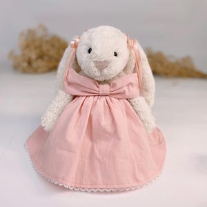 May include: A white plush bunny wearing a pink dress with a bow and lace trim. The bunny has pink ribbons in its ears.