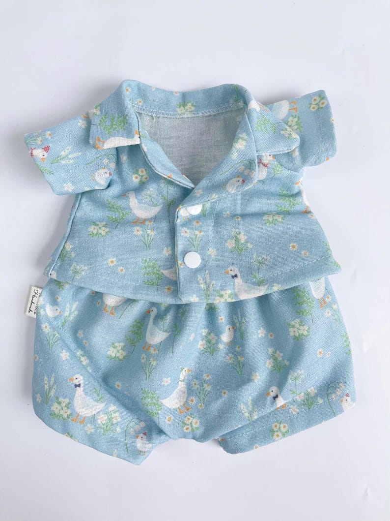 Puede incluir: Un conjunto de mu&ntilde;eca beb&eacute; azul claro con una camisa de manga corta y pantalones cortos. La tela tiene un patr&oacute;n blanco y verde de patos y flores.