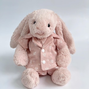 Puede incluir: Un conejito de peluche rosa con un pijama rosa y blanco estampado. El conejito tiene una expresi&oacute;n dulce y est&aacute; sentado sobre un fondo blanco.