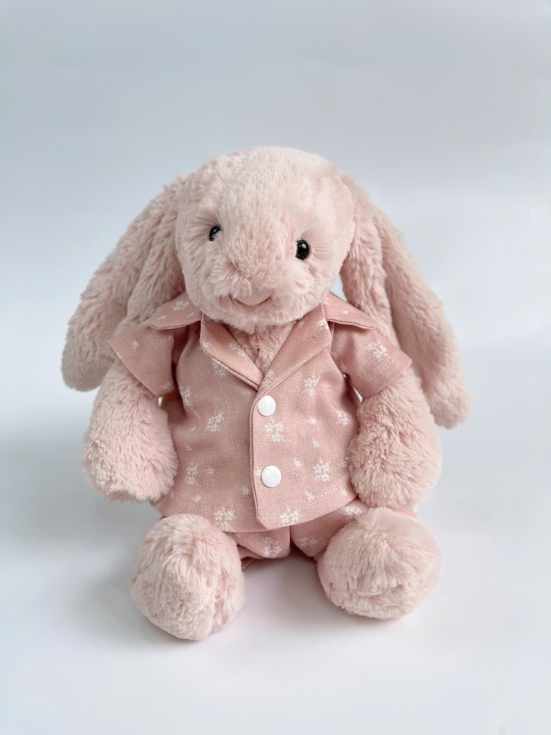 Puede incluir: Un conejito de peluche rosa con un pijama rosa y blanco estampado. El conejito tiene una expresi&oacute;n dulce y est&aacute; sentado sobre un fondo blanco.