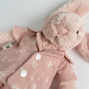 Puede incluir: Un conejito de peluche rosa con un pijama rosa de estampado floral blanco con botones blancos.