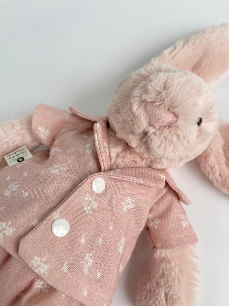 Puede incluir: Un conejito de peluche rosa con un pijama rosa de estampado floral blanco con botones blancos.