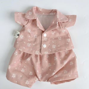 Puede incluir: Un conjunto de pijama rosa con estampado floral blanco para una mu&ntilde;eca. El conjunto incluye una camisa de manga corta con botones en la parte delantera y pantalones cortos a juego.