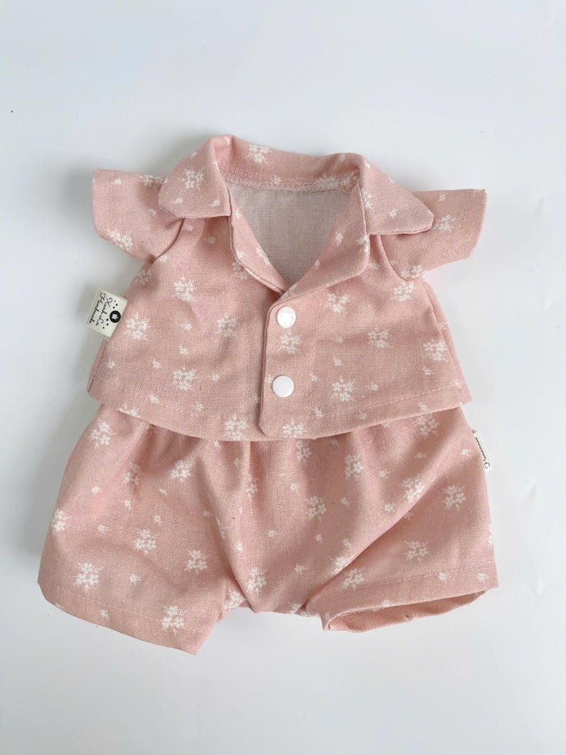 Puede incluir: Un conjunto de pijama rosa con estampado floral blanco para una mu&ntilde;eca. El conjunto incluye una camisa de manga corta con botones en la parte delantera y pantalones cortos a juego.