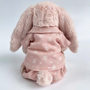 Puede incluir: Un conejito de peluche rosa con un pijama rosa de estampado floral blanco. El conejito tiene orejas largas y ca&iacute;das y una cola esponjosa.
