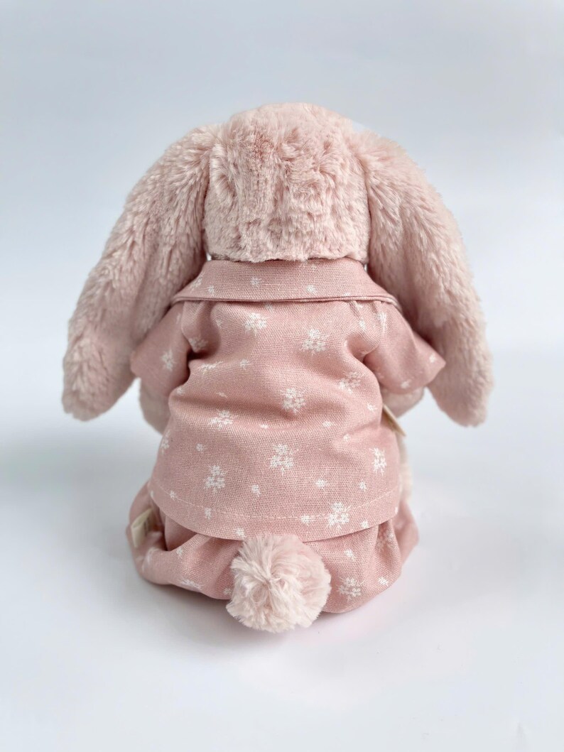 Puede incluir: Un conejito de peluche rosa con un pijama rosa de estampado floral blanco. El conejito tiene orejas largas y ca&iacute;das y una cola esponjosa.