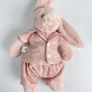 Puede incluir: Un conejito de peluche rosa con un pijama rosa de estampado floral blanco. El conejito tiene orejas largas y una expresi&oacute;n dulce.
