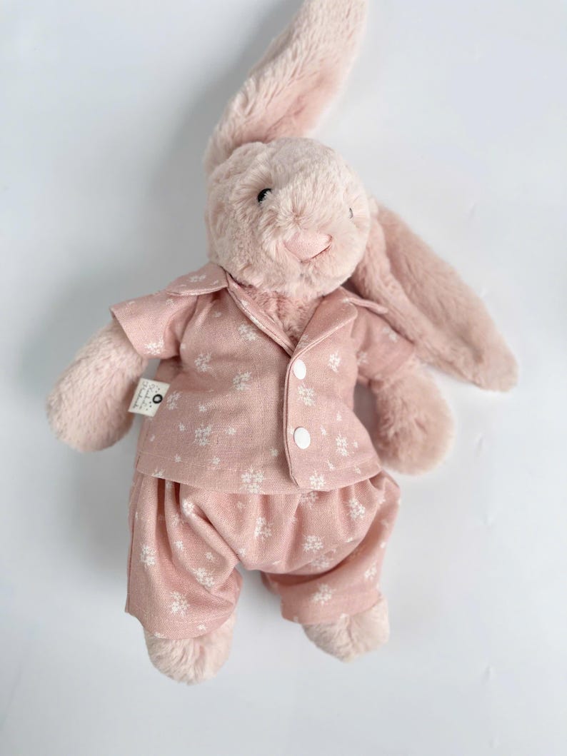Puede incluir: Un conejito de peluche rosa con un pijama rosa de estampado floral blanco. El conejito tiene orejas largas y una expresi&oacute;n dulce.