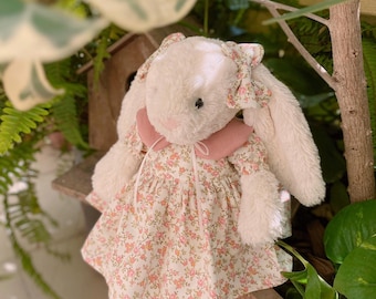 May include: A plush white bunny wearing a floral print dress with a pink collar and bow. The bunny has long, floppy ears and is positioned in front of greenery. The dress features a pattern of small pink and orange flowers.