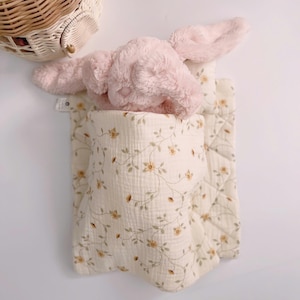 Puede incluir: Un juguete de peluche rosa en forma de conejo con orejas largas está acostado en una manta de estampado floral blanco y verde. La manta tiene un borde rizado.