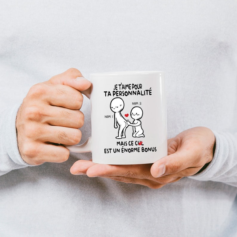 May include: White ceramic mug with a black and white illustration of two stick figures. The text on the mug reads "JE T'AIME POUR TA PERSONNALIT&Eacute; MAIS CE CUL EST UN &Eacute;NORME BONUS".