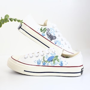 Könnte beinhalten: Ein Paar weiße Canvas-Sneaker mit niedrigem Schaft und einem blauen, grünen und weißen Meereslebewesen-Design. Das Design umfasst eine Schildkröte, eine Robbe, Algen und Blasen.