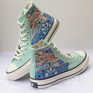 May include: A pair of mint green high-top trainers with mosaic-style artwork on the sides. The trainers have white laces, a white toe cap, and a black stripe on the sole. The artwork features blue, white, and other colourful geometric shapes.