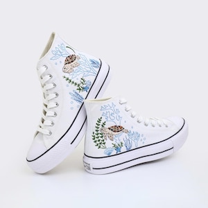 May include: A pair of white high-top sneakers featuring embroidered sea turtle designs on the outer sides. The embroidery includes light blue coral, green seaweed, and small seashells. The shoes have white laces, white rubber soles with black trim.
