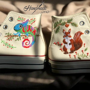 May include: A pair of white high-top canvas shoes with embroidered designs. One shoe features a colorful chameleon on a branch with red berries, while the other shoe has a brown squirrel with a bushy tail surrounded by green leaves and red mushrooms.