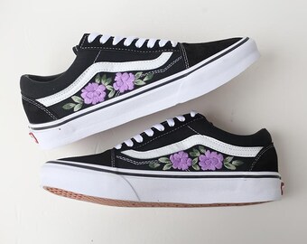 May include: Black sneakers with white soles and laces. The shoes feature a white side stripe and embroidered purple floral designs. The soles are white with a brown bottom. The shoes are a classic style.