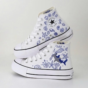 May include: White high-top sneakers with blue seashell and shark designs. The sneakers feature a black stripe on the sole and the Converse All Star logo. The design includes various sea creatures and marine elements.